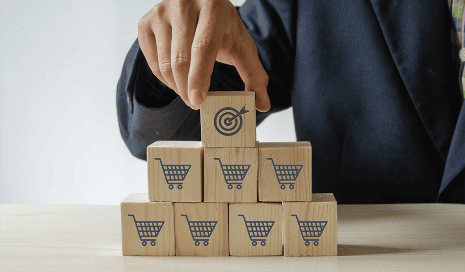 Homem empilha cubos de madeira com marcas de carrinhos de compra e no topo um cubo com desenho de alvo representando as metas de vendas.
