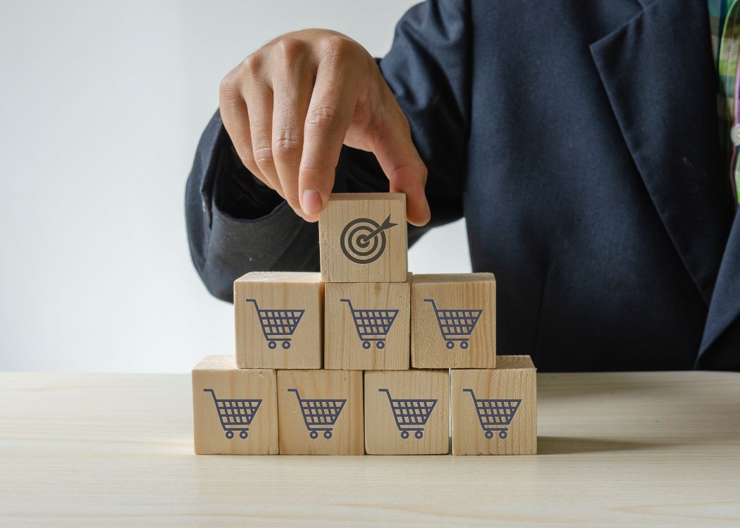 Homem empilha cubos de madeira com marcas de carrinhos de compra e no topo um cubo com desenho de alvo representando as metas de vendas.